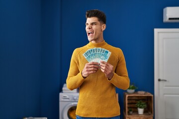 Young hispanic man at laundry room holding brazilian reals angry and mad screaming frustrated and furious, shouting with anger. rage and aggressive concept.