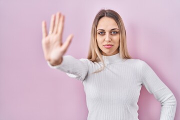 Young blonde woman wearing white sweater over pink background doing stop sing with palm of the hand. warning expression with negative and serious gesture on the face.