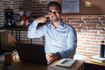 Hispanic man with beard working at the office at night pointing with hand finger to face and nose, smiling cheerful. beauty concept