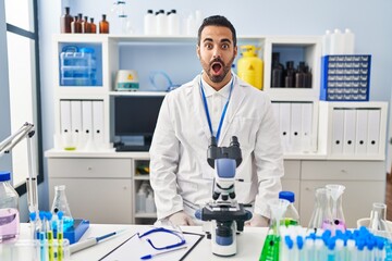 Fototapeta premium Young hispanic man with beard working at scientist laboratory afraid and shocked with surprise expression, fear and excited face.