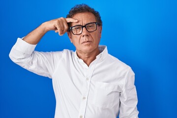 Naklejka premium Middle age hispanic man standing over blue background pointing unhappy to pimple on forehead, ugly infection of blackhead. acne and skin problem