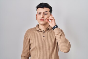 Non binary person standing over isolated background mouth and lips shut as zip with fingers. secret...