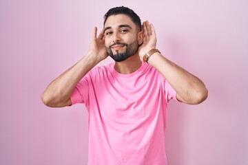 Hispanic young man standing over pink background trying to hear both hands on ear gesture, curious for gossip. hearing problem, deaf