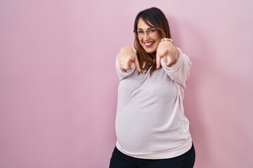 Pregnant woman standing over pink background pointing to you and the camera with fingers, smiling positive and cheerful