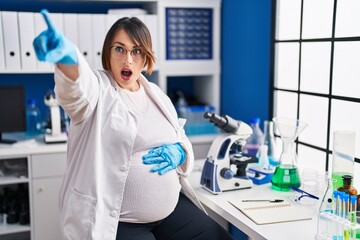 Pregnant woman working at scientist laboratory pointing with finger surprised ahead, open mouth...