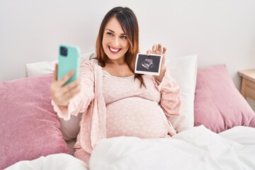 Young pregnant woman make selfie by smartphone holding ecography at bedroom