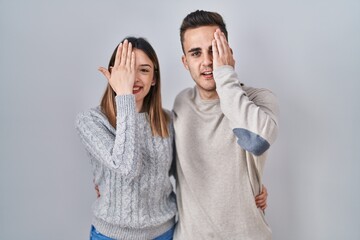 Young hispanic couple standing over white background covering one eye with hand, confident smile on face and surprise emotion.