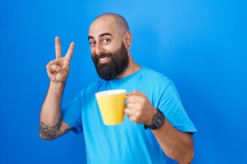 Young hispanic man with beard and tattoos drinking a cup of coffee smiling with happy face winking at the camera doing victory sign with fingers. number two.