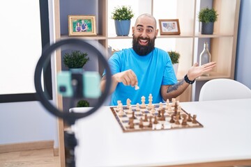 Young hispanic man with beard and tattoos recording chess tutorial with smartphone at home...