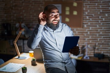 Young hispanic man with beard and tattoos working at the office at night smiling with hand over ear listening an hearing to rumor or gossip. deafness concept.