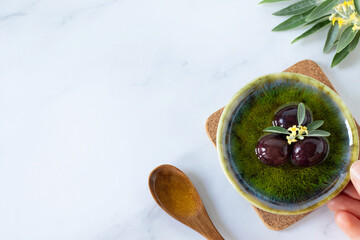 Hand holding bowl with natural extra virgin olive oil and black olives, green tree branch and...