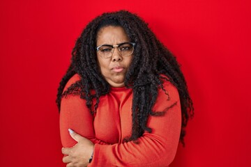 Plus size hispanic woman standing over red background shaking and freezing for winter cold with sad and shock expression on face