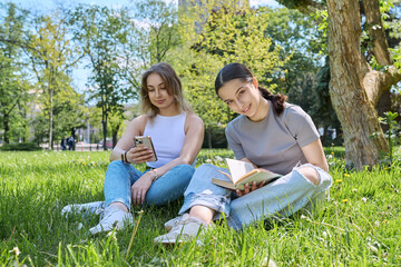 Fototapeta premium Two teenage girls friends relaxing on grass using smartphone reading book