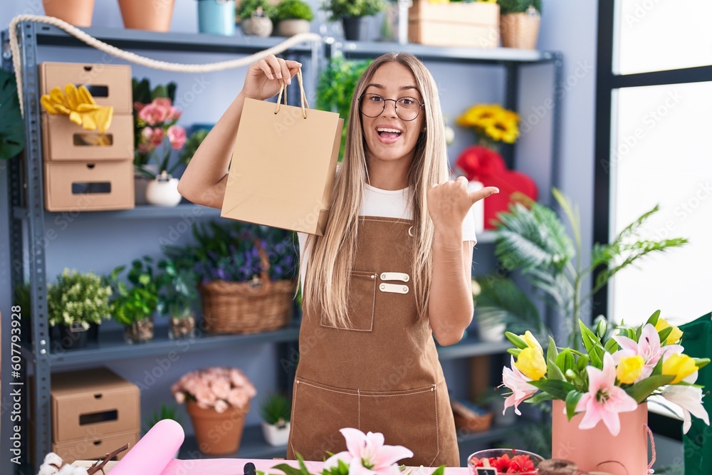 Wall mural Young blonde woman working at florist shop holding bag pointing thumb up to the side smiling happy with open mouth