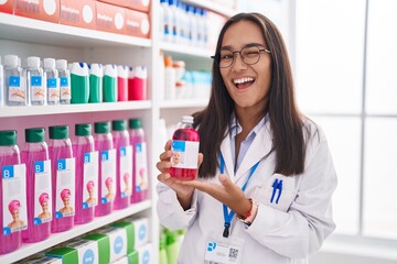 Young hispanic woman working at pharmacy drugstore holding syrup winking looking at the camera with...