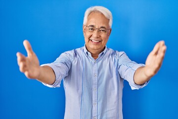 Hispanic senior man wearing glasses looking at the camera smiling with open arms for hug. cheerful expression embracing happiness.