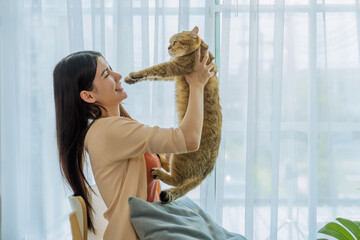 Cherished moment of a young Asian woman lifting her adorable cat to her face by the bright morning...