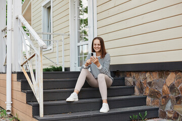 Obraz premium Portrait of smiling attractive caucasian woman wearing striped shirt using phone sitting on porch of house browsing internet reading messages chatting with friends.