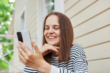 Smiling happy woman wearing striped shirt using phone posing near house expressing positive emotions watching funny videos on smartphone having video call.