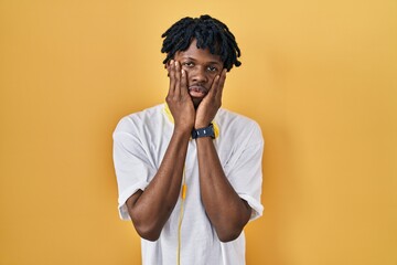 Young african man with dreadlocks standing over yellow background tired hands covering face, depression and sadness, upset and irritated for problem