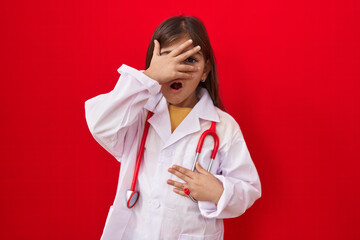 Little hispanic girl wearing doctor uniform and stethoscope peeking in shock covering face and eyes...