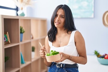 Brunette woman eating a salad at home clueless and confused expression. doubt concept.