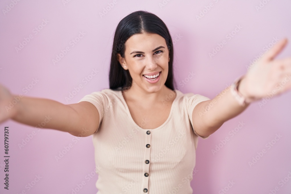 Poster young hispanic woman standing over pink background looking at the camera smiling with open arms for 