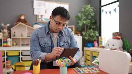 Young chinese man preschool teacher using touchpad sitting on table at kindergarten