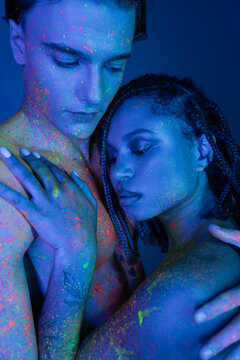 sensual interracial couple in colorful body paint embracing on blue background with cyan lighting, handsome bare-chested man and enchanting african american woman with dreadlocks