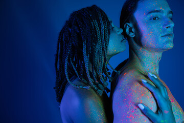 intimate moment of youthful interracial couple in colorful neon body paint on blue background with...