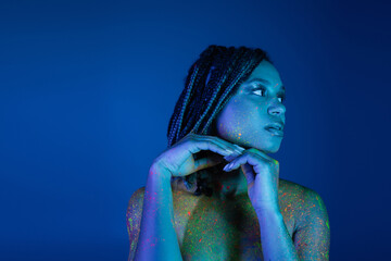 young and captivating african american woman with dreadlocks, in bright and colorful neon body paint holding hands near chin and looking away on blue background with cyan lighting effect
