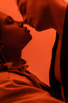 side view of young and romantic interracial couple in love, charismatic man and sensual african american woman kissing with closed eyes on orange background with red lighting effect