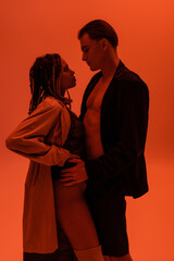 side view of sexy man in black blazer on shirtless body hugging provocative african american woman in lace bodysuit and beige trench cost on orange background with red lighting effect