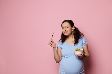 Adorable multi ethnic pregnant woman holding a bowl with fresh organic fruits, enjoying healthy lifestyle and wholesome nutrition in pregnancy time, smiling at camera, over pink background. Copy space