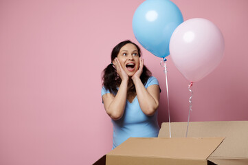 Emotional portrait of pregnant woman, expressing happy surprise while two balloons appears from a box, feeling excitement expecting twins, isolated on pink background. Gender reveal party. Baby shower