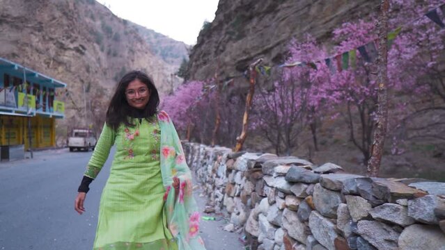 Beautiful Indian girl wearing Traditional ethnic Indian dress walking on the road besides flowering apricot trees and mountains in background at Kinnaur, India. Indian ethnic woman outdoors.
