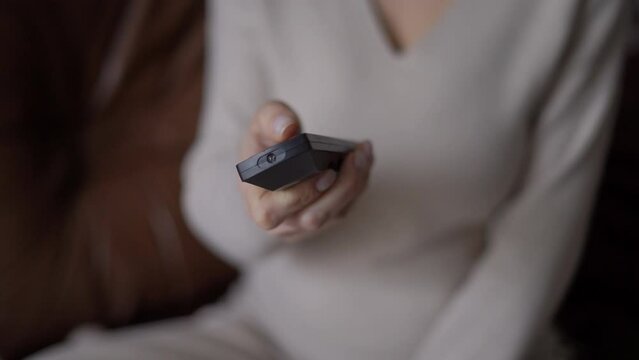 Closeup remote control in hand of pregnant unrecognizable woman switching channels on TV. Young relaxed Caucasian expectant enjoying leisure at home indoors watching movies