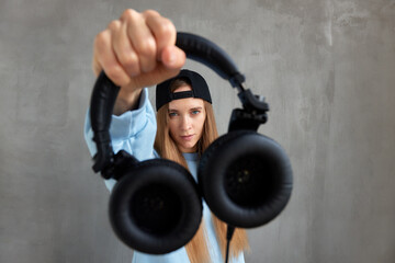 A young pretty long-haired DJ girl in a blue sweater and a funny blue hat holds black headphones in outstretched hands. Studio shot, gray background.
