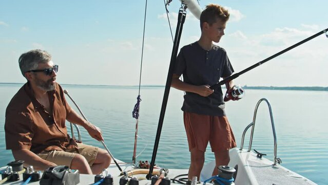 Mature father watching his teen son fishing in lake using rod during trip on sailboat on sunny summer day