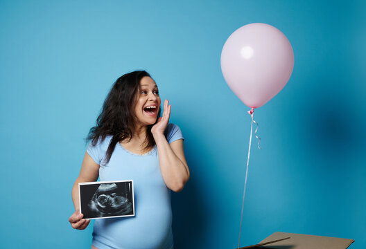 Pregnant woman experiencing joy while watching a pink balloon flying out of a cardboard box, smiling, holding ultrasound scan image of her future child, isolated on blue backdrop. Gender reveal party