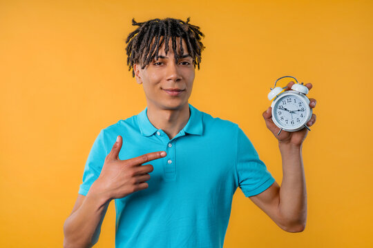 Happy student african man with alarm clock on yellow backdrop.