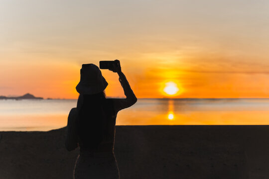 Silhouette of woman with mobile phone taking a photo of the sunset on the beach.