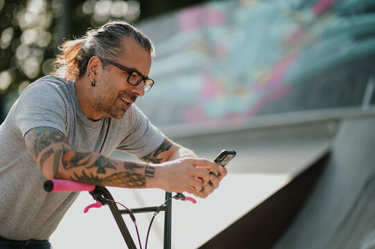 Portrait of a happy urban middle-aged guy leaning on his bmx bike in the skate park and using his phone for telecommunications.