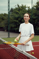 Vertical photo of young attractive girl standing near tennis net at tennis court with racket and ball in hands