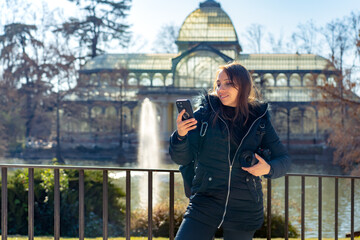 Happy young Latin woman in warm clothes with professional photo camera standing near using smartphone in front of Glass Palace in Madrid, Spain