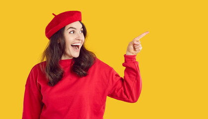 Happy excited attractive young brunette woman in red beret and sweatshirt standing on yellow studio background, looking aside and pointing index finger at copy space side. Autumn fashion sale concept