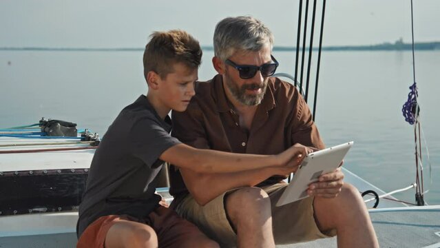Mature Caucasian man and his teen son sitting on board the yacht using reading map or surfing Internet in digital tablet