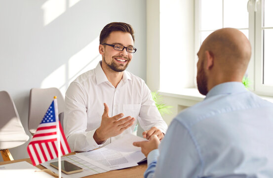 Happy, smiling, friendly young male Consul of United States of America sitting at office table with American flag and talking to man about his immigration plans and USA travel visa application
