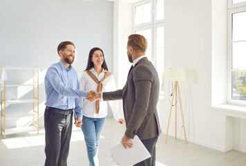 Happy couple buying house. Man and woman standing in modern white light living room and shaking hands with real estate agent. Young family buy property, make deal and exchange handshake with relator - Powered by Adobe