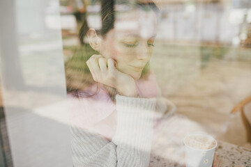 Young woman sitting near window in a coffeehouse, enjoying latte.